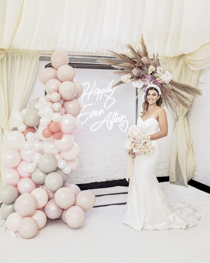 Bridal model posing in front of a balloon stand with a neon Happily Ever After sign and floral and feather display.